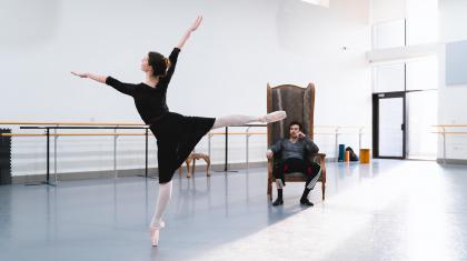 A dancer stood on pointe with legs and arms outstretched with another dancer watching on in a large armchair