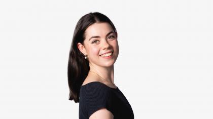 A dancer stood on a white background with medium length brown hair smiling to camera