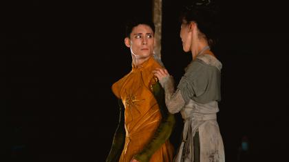 Merlin, wearing a golden tunic, looks worriedly at his adoptive mother, The Blacksmith