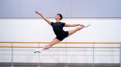 Dancer leaping across the frame in a black T-shirt and shorts, looking powerful as he stretches is legs and arms in front and behind.
