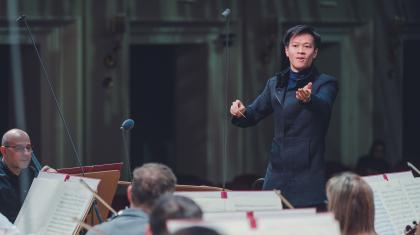 Conductor wearing black, left hand raised to the musicians, right hand holding the baton